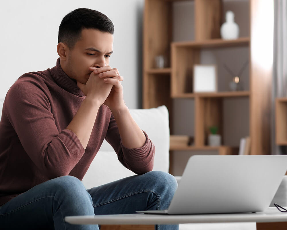 Photo of a man sitting on his couch looking at his laptop with his hands folded together covering his mouth. Have your anxiety symptoms taken over your daily routine? Learn how anxiety therapy in Atlanta, GA can help you manage your anxiety symptoms with coping skills like meditation skills.