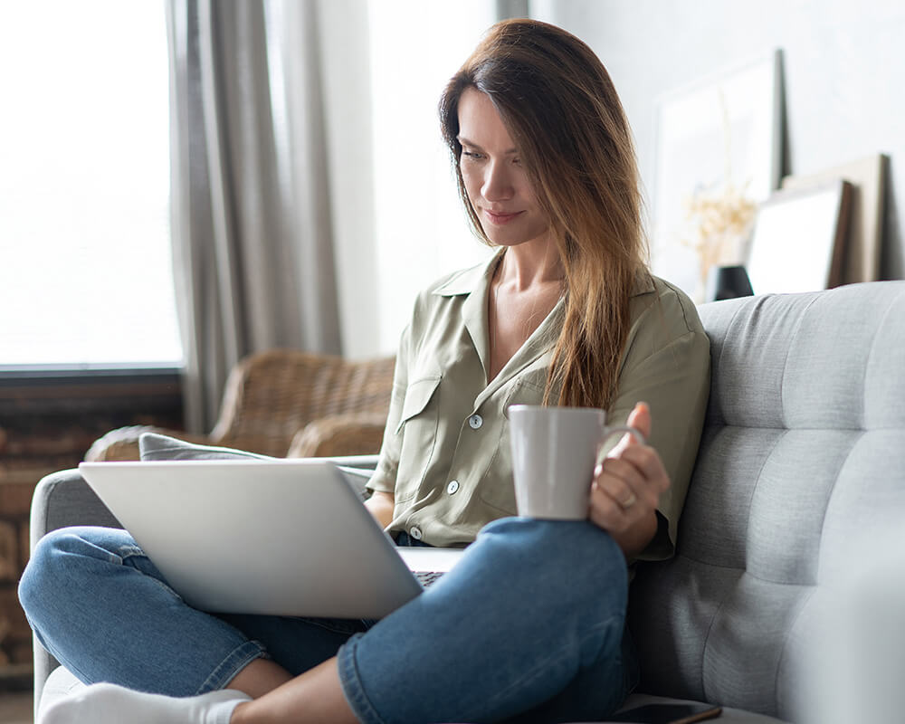 Photo of a woman sitting on her couch using her laptop while drinking out of a mug. This photo represents how easy it is to receive depression counseling in Atlanta, GA. Learn more here!