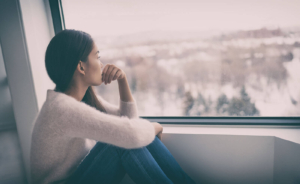 Photo of a sad woman sitting on the ground looking out the window. This photo represents how grief can be overwhelming and how grief counseling in Atlanta, GA can help you begin to cope with your grief symptoms.