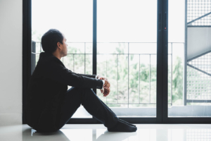 Photo of a sad man sitting on the floor holding his knees and looking out a window. With grief counseling in Atlanta, GA you can begin to process your emotions and learn to manage your grief more effectively.