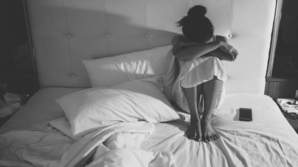 Black and white photo of a woman sitting in bed hugging her knees. If you are struggling with isolation due to your depression don't wait to get help. With depression therapy in Atlanta, GA you can begin your journey to start healing from your symptoms.