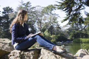 Photo of a woman sitting on a rock outside looking at a pond and journaling. This photo represents how journaling can help in identifying your stress triggers. Discover how therapy for stress management in Atlanta, GA can help you begin managing your symptoms.