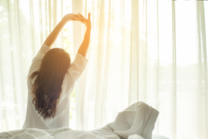 Photo of a woman sitting on her bed stretching and waking up. Discover tips on staying motivated while working from home. Learn more tips on motivation and focusing without stress with stress therapy in Atlanta, GA.