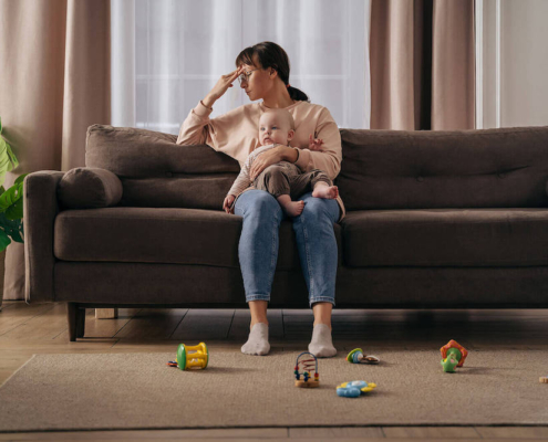 Image of an upset mom sitting on a couch with her hand on her forehead and holding a baby. Learn how to deal traumatic births with the help of birth trauma therapy. Find support in postpartum therapy in Atlanta, GA.