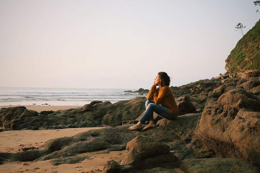 Photo of a woman sitting on a rocky beach looking out at the ocean. Discover the first step to controlling your anxious thoughts and how anxiety therapy in Atlanta, GA can help you begin challenging those thoughts.