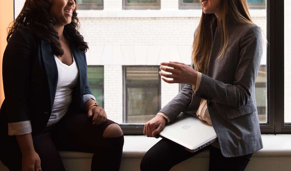 Two professional women sitting on a windowsill smiling and speaking with each other. Find help managing your anxious thoughts with the help of anxiety therapy in Atlanta, GA.