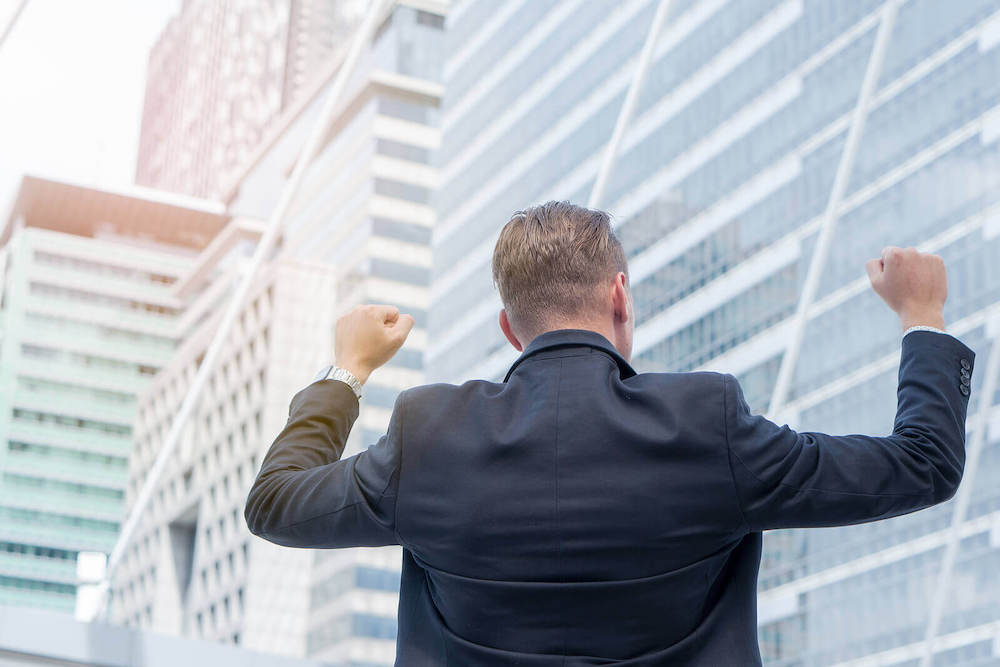 Image of a business man wearing a suit lifting his arms in victory while standing in front of a skyscraper. Overcome your workplace anxiety and create a healthy work life balance with the help of an online anxiety therapist in anxiety therapy in Atlanta, GA. Find relief soon.