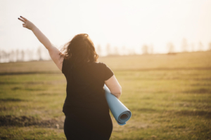 A woman holding a yoga mat walks across a field while holding up a peace sign. This could represent the benefits of working with a postpartum therapist in Atlanta, GA to address depression. Search for online depression therapy in Atlanta, GA and other services by searching for depression therapy in Atlanta, GA today. 