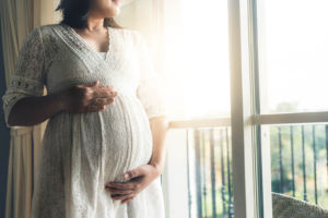 A woman holds her pregnant stomach while looking out a window. Learn how a postpartum therapist in Atlanta, GA can offer support via online depression therapy in Atlanta, GA. Contact an online depression therapist in Atlanta, GA for support. 