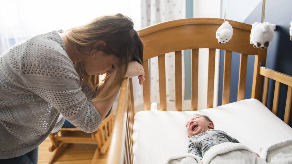 A mother rests her head on the side of her crying baby's crib. Learn how postpartum therapy in Atlanta, GA can help address anxiety. Contact an online anxiety therapist in Atlanta, GA for support with therapy for anxiety in Atlanta, GA and more.