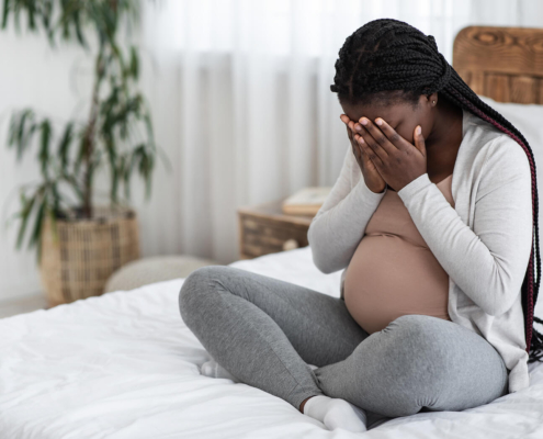 A pregnant woman covers her face while sitting on a bed. This could represent the postpartum pain that a postpartum therapist in Atlanta, GA can address. Learn more about therapy for birth trauma in Atlanta, GA by searching for "postpartum therapist atlanta" today.