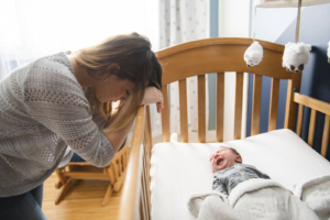 A mother rests her head on a crib of her crying baby. This could represent the stress of postpartum that a depression therapist in Atlanta, GA can address. Learn how a postpartum therapist in Atlanta, GA can help you by searching for birth trauma therapy in Atlanta, GA today. 