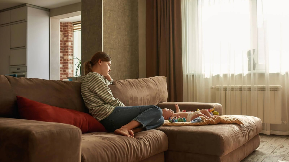A tired mom sits on a couch while looking at her tired baby. This could represent the support that therapy for birth trauma in Atlanta, GA can offer in addressing postpartum issues. Learn more about postpartum therapy in Atlanta, GA by contacting a postpartum therapist in Atlanta, GA today.