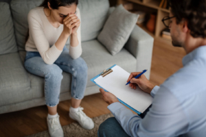 A man holds a notepad while sitting across from a woman bowing her head with a concerned expression. This could represent the support an anxiety therapist in Atlanta, GA can offer via stress and anxiety counseling in Atlanta, GA. Search for anxiety therapy in Atlanta, GA today. 