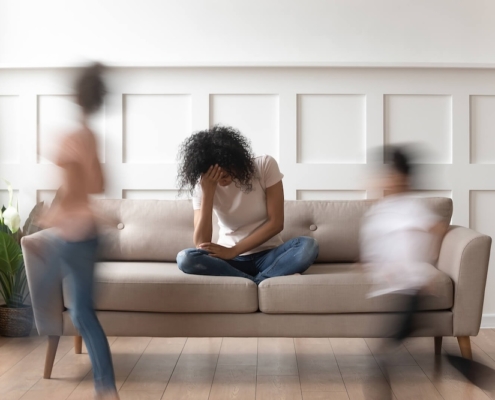 Image of an overwhelmed mom sitting on a couch while her children run. Discover how postpartum therapy in Atlanta, GA can help you navigate postpartum life.
