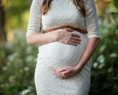 Close-up of a pregnant woman in a white lace dress gently holding her stomach in a serene outdoor setting, symbolizing pregnancy therapy in Atlanta, GA, and the compassionate care of a perinatal therapist in Atlanta, GA for those navigating grief & loss therapy in Atlanta.