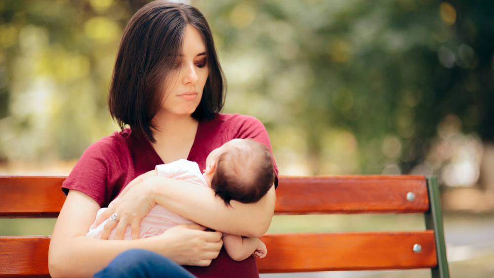 A new mother sits on a park bench holding her baby, appearing overwhelmed—symbolizing the emotional weight that postpartum therapy in Atlanta, GA or postpartum anxiety treatment in Atlanta can help address.