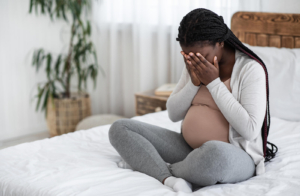A pregnant woman sits on her bed with her hands covering her face, showing the struggle of grief and worry while finding postpartum therapy in Atlanta, GA, anxiety therapy in Atlanta, GA, postpartum counseling Atlanta, and Alpharetta postpartum anxiety treatment. 
