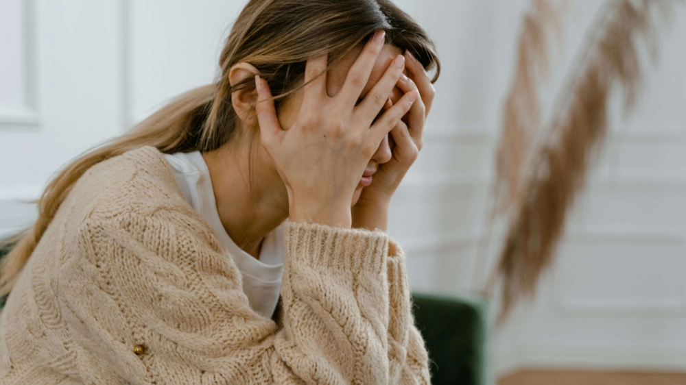 A young woman sits on a couch with her hands covering her face, visibly overwhelmed and distressed. This emotional moment may reflect the experience of postpartum rage in Atlanta, GA — a common but often overlooked response in the postpartum period. Support from a postpartum therapist in Atlanta, GA can help those facing intense emotional shifts. Alpharetta postpartum anxiety treatment offers a safe space to process these difficult feelings.