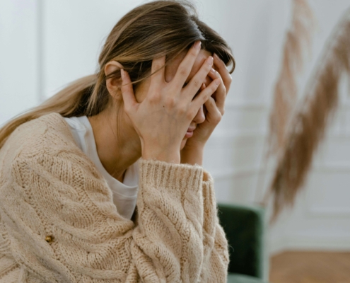 A young woman sits on a couch with her hands covering her face, visibly overwhelmed and distressed. This emotional moment may reflect the experience of postpartum rage in Atlanta, GA — a common but often overlooked response in the postpartum period. Support from a postpartum therapist in Atlanta, GA can help those facing intense emotional shifts. Alpharetta postpartum anxiety treatment offers a safe space to process these difficult feelings.